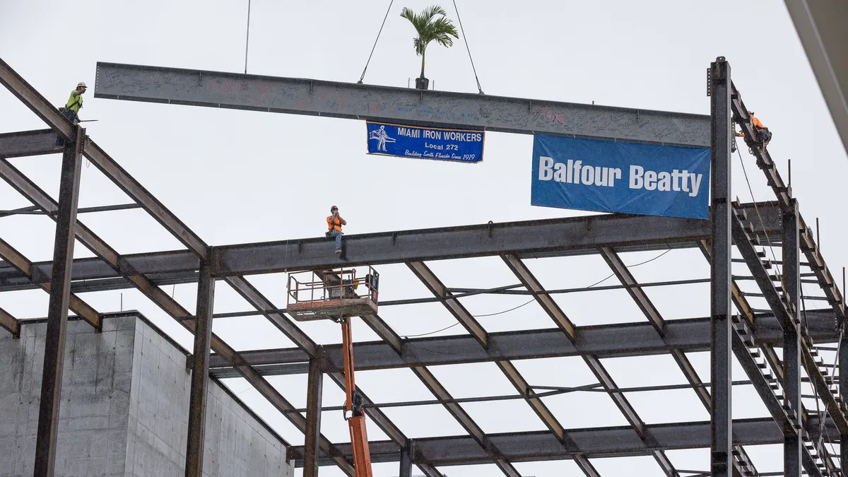 A person wearing safety gear directs a beam that has a flag of the company Balfour Beatty, along with a Miami ironworkers union flag.