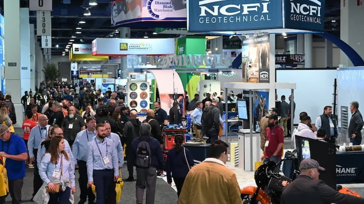 A large group of people in clothes ranging from casual to business casual mill about an exhibit hall at a trade conference. Booths are set up all around them, and people mill about.