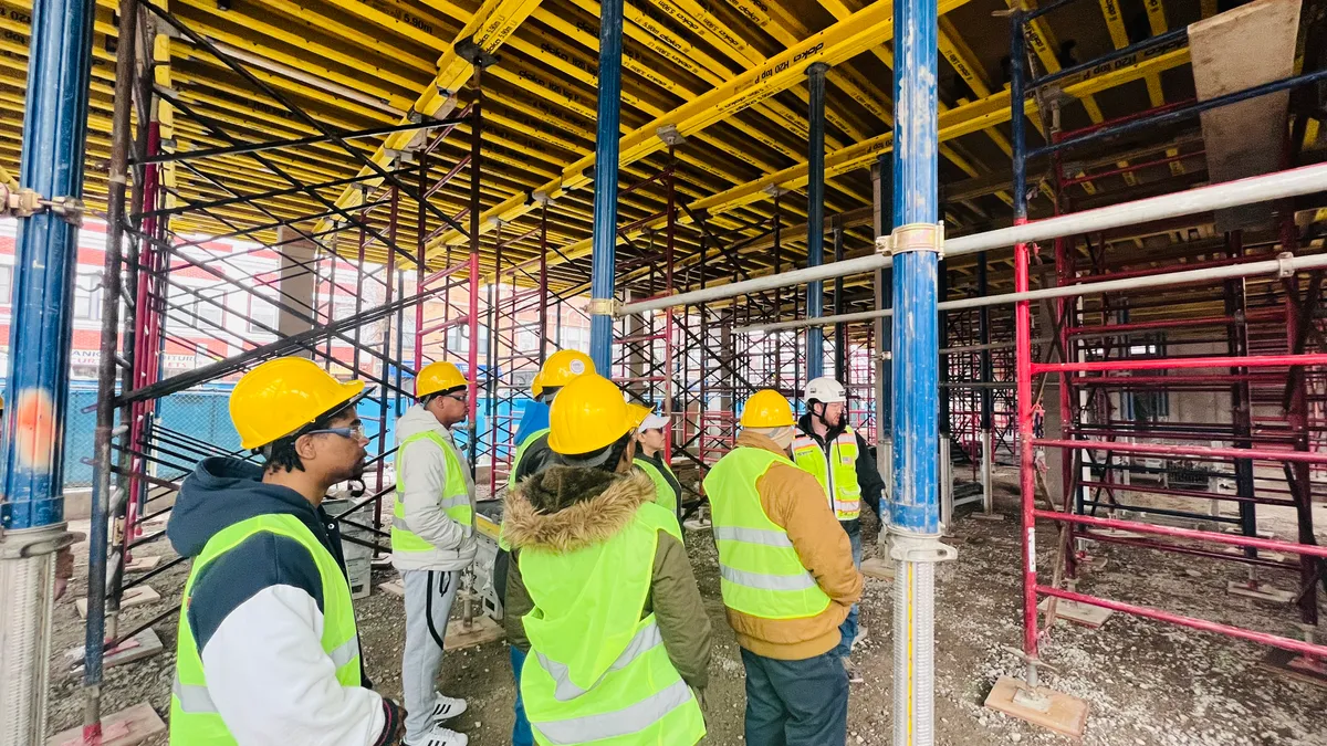 A group of people in hard hats and yellow vests walk between scaffolding on a jobsite.